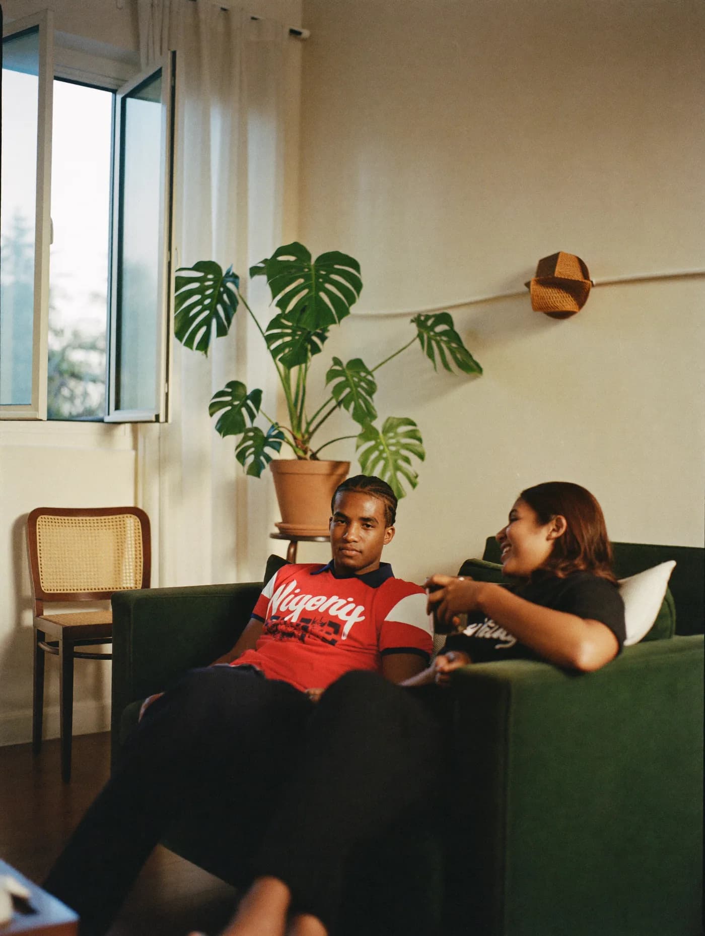 Two housemates talking on a green sofa in a sunlit Lagos flat.